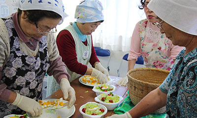 三角巾をかぶり、華やかなエプロンをかけたおばさんたちが、サラダをつくっています。